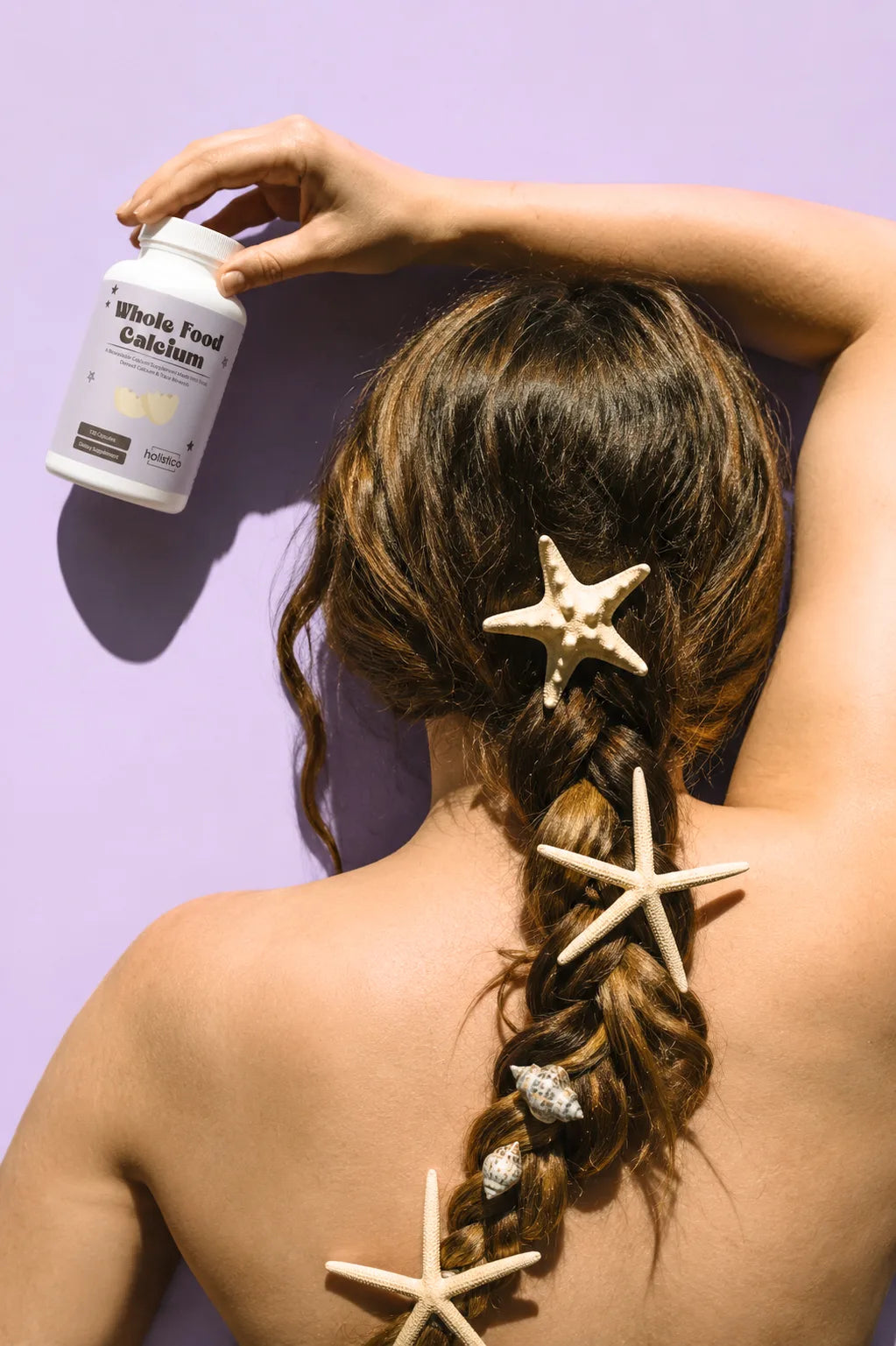 A hand placing a bottle of Holistico Whole Food Calcium against a lilac background, hair braided with starfish and shells