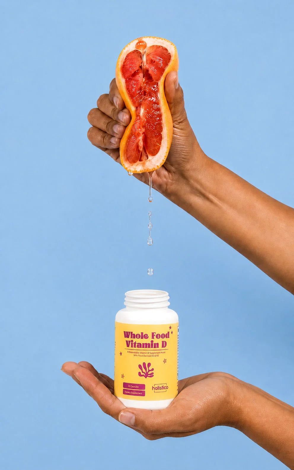 A hand squeezing grapefruit juice into a bottle of Holistico Whole Food Vitamin D against a blue background