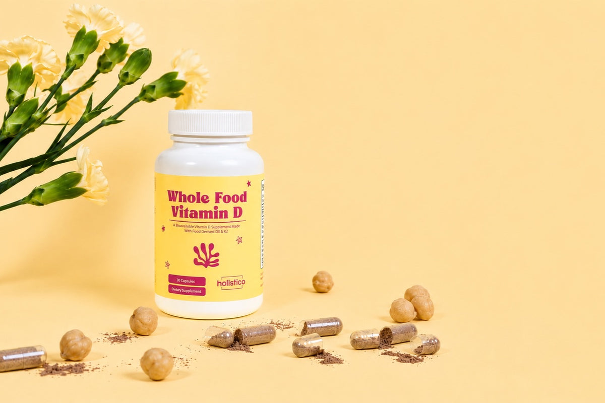A bottle of Holistico Whole Food Vitamin D beside yellow flowers, chickpeas and open capsules on a butter yellow ground