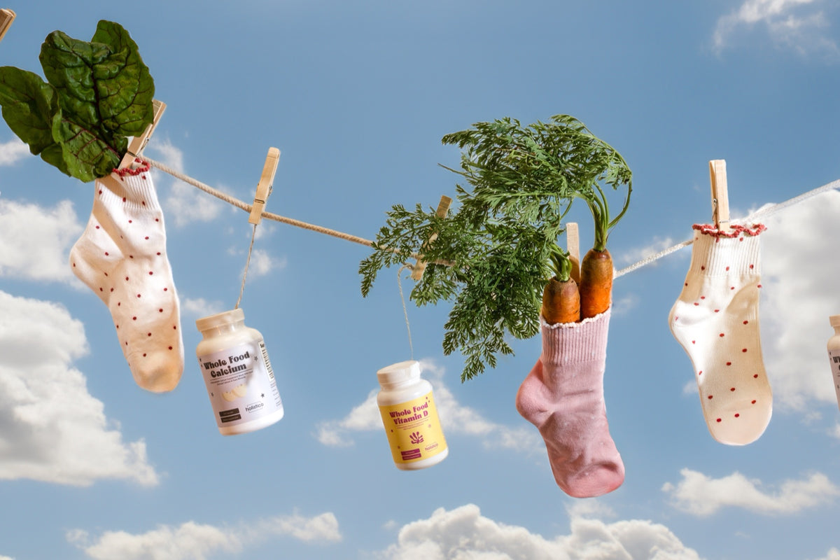 Holistico Whole Food Vitamin D and Whole Food Calcium bottles pegged to a clothesline with carrots and leafy greens against a sunny sky