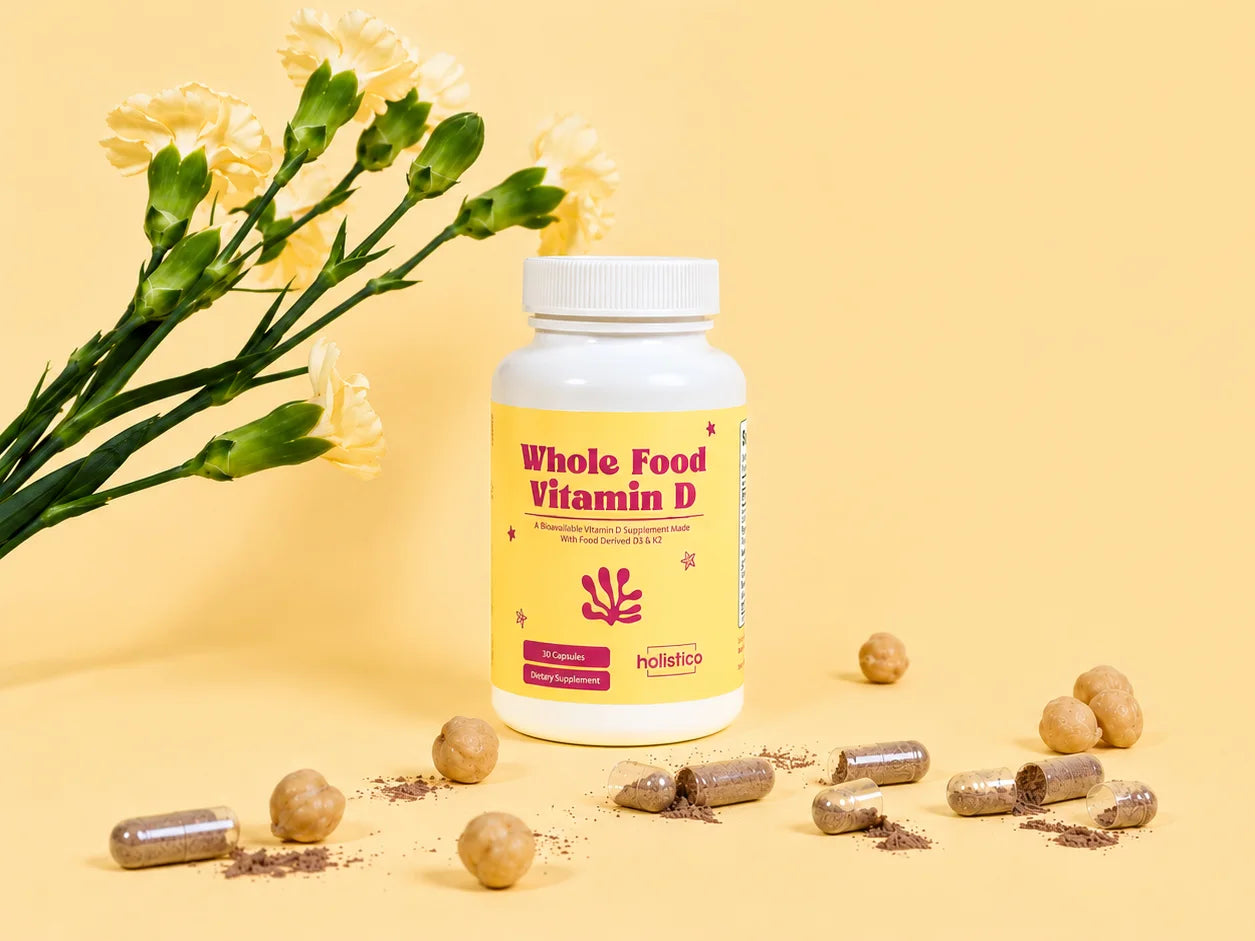 A bottle of Holistico Whole Food Vitamin D beside a bunch of pale yellow carnations, with capsules scattered on a butter yellow background