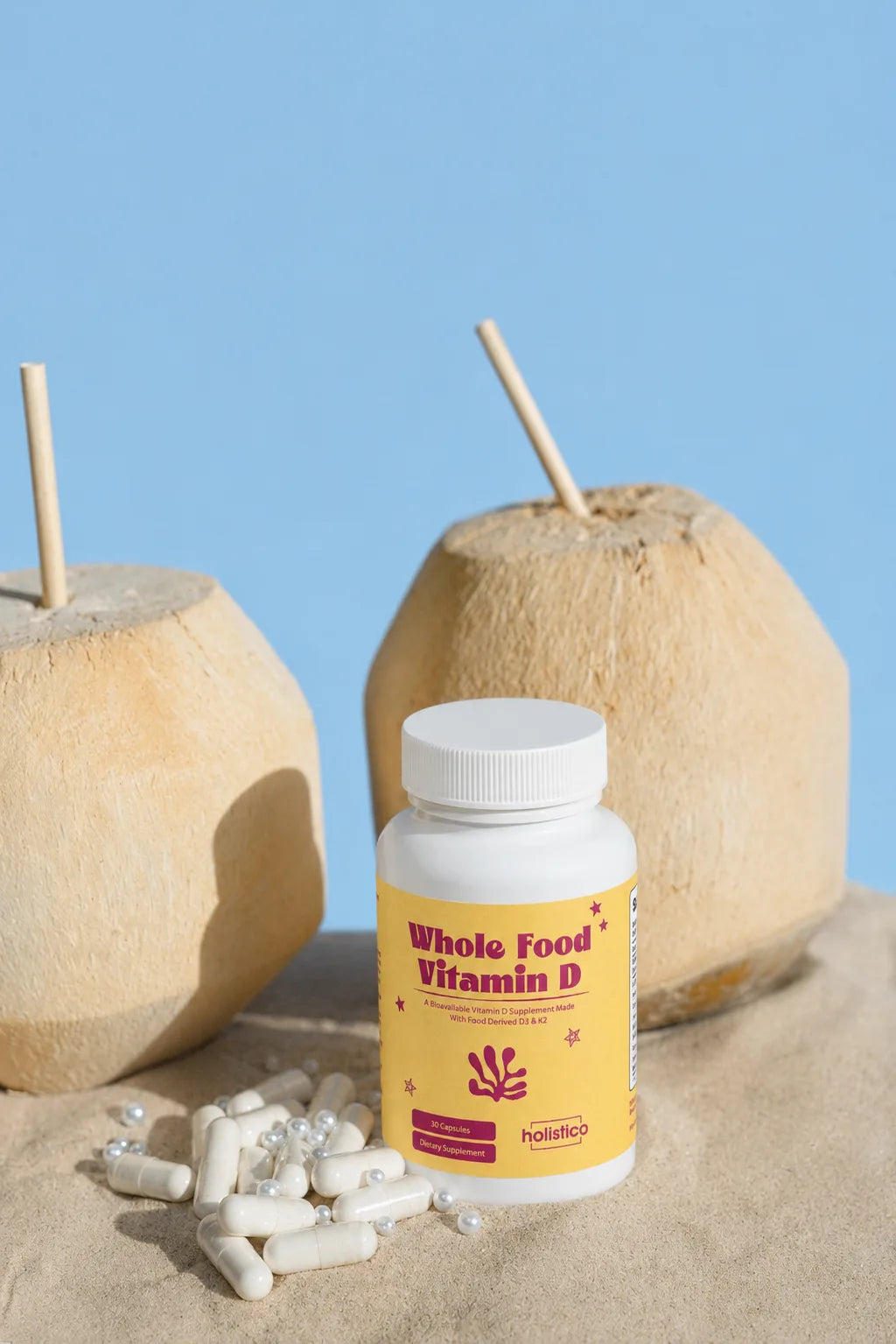 A bottle of Holistico Whole Food Vitamin D standing in sand between two whole coconuts with straws, against a blue background