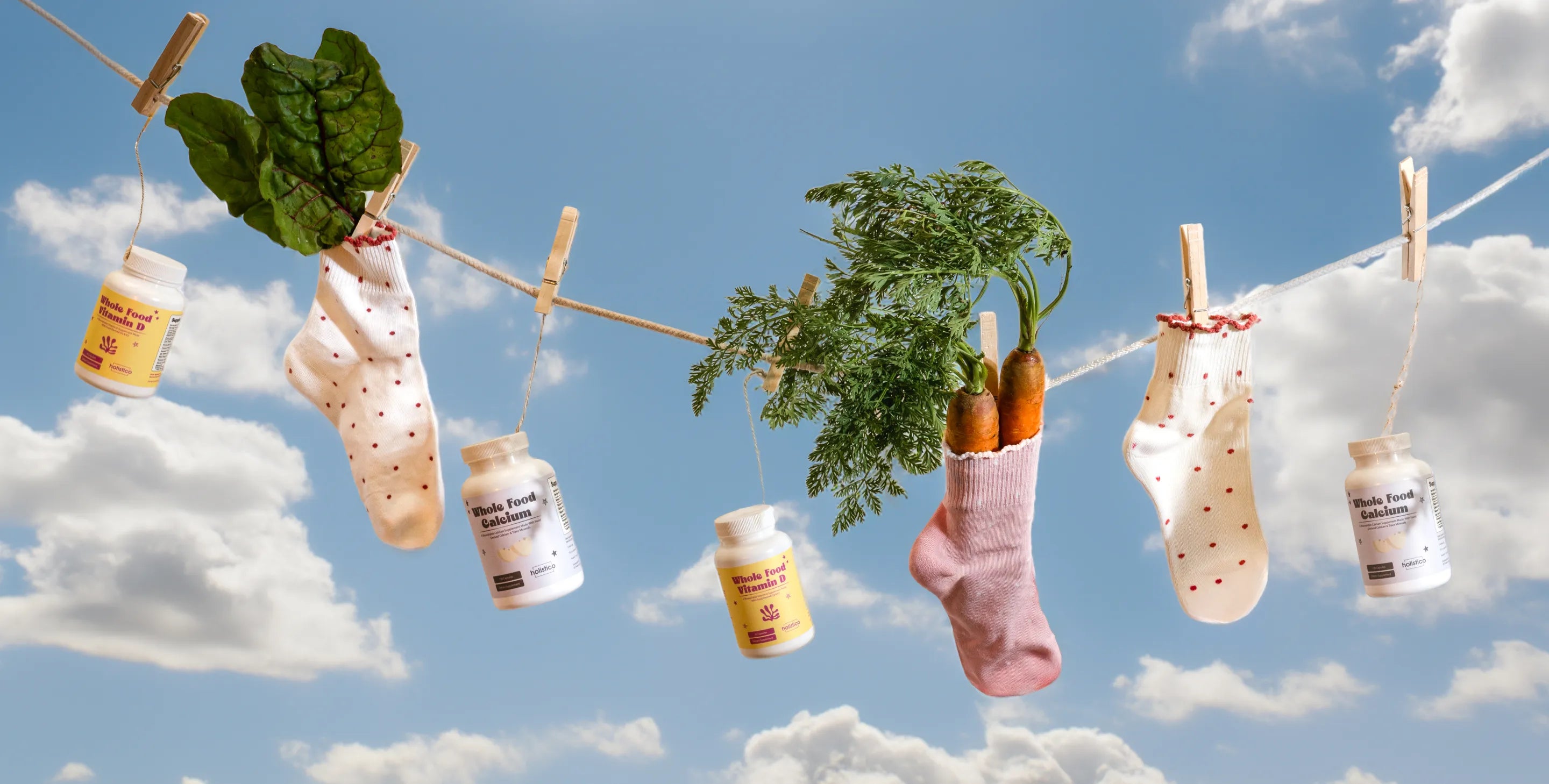 Bottles of Holistico Whole Food Calcium and Whole Food Vitamin D pegged to an outdoor clothesline against a blue sky, alongside socks, chard leaves and carrots