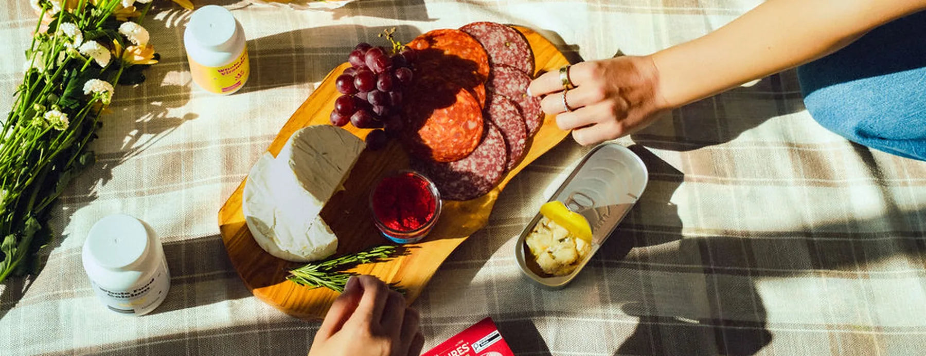 An overhead picnic spread on a tartan blanket -- a charcuterie board, flowers and two bottles of Holistico Whole Food Vitamin D and Whole Food Calcium