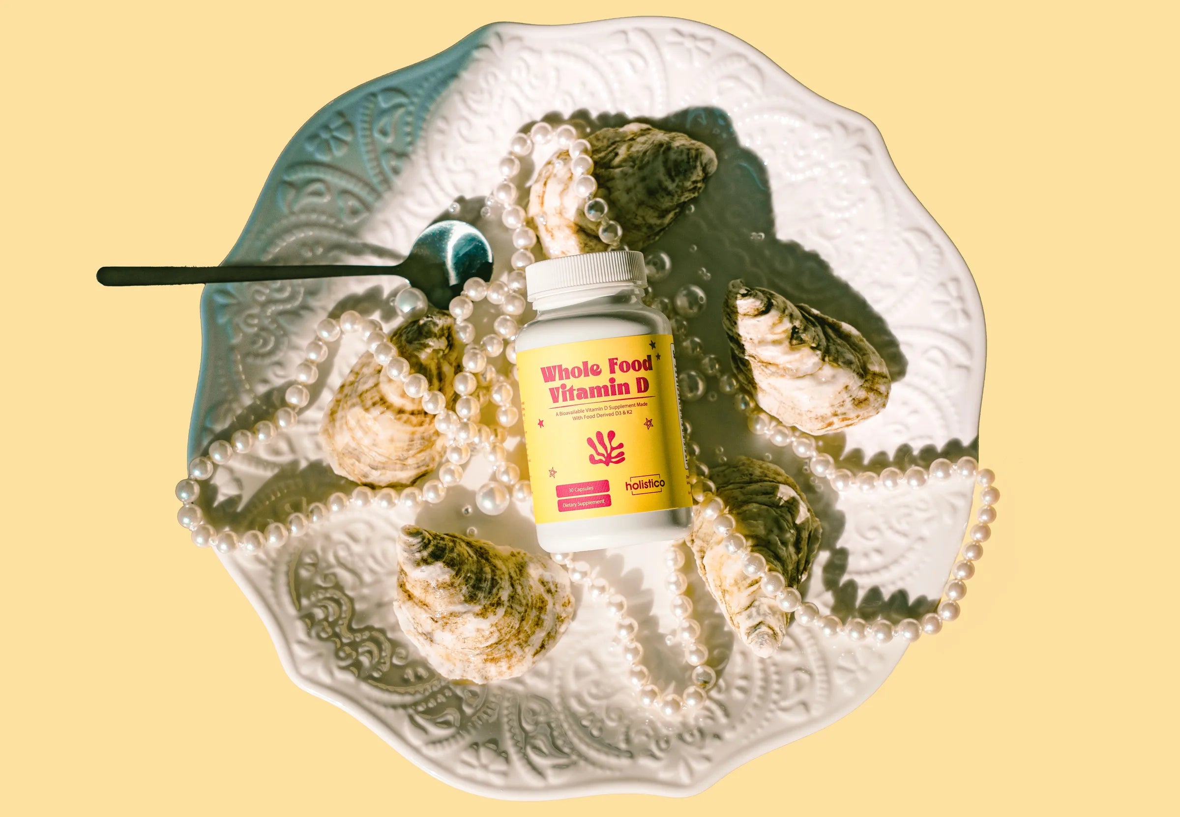 A bottle of Holistico Whole Food Vitamin D on an ornate white plate with oysters and strings of pearls, on a yellow background