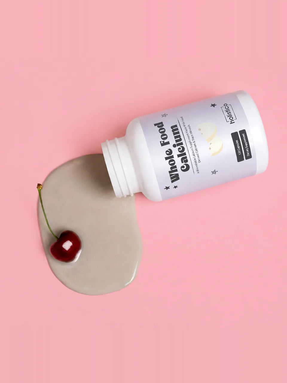 A bottle of Whole Food Calcium tipped on its side on a pink background, a creamy pour spilling out around a single cherry