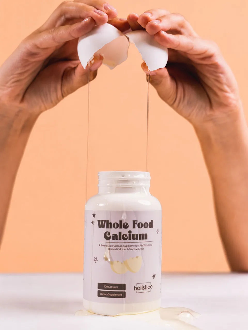 Two hands cracking an eggshell above an open bottle of Whole Food Calcium on a peach background