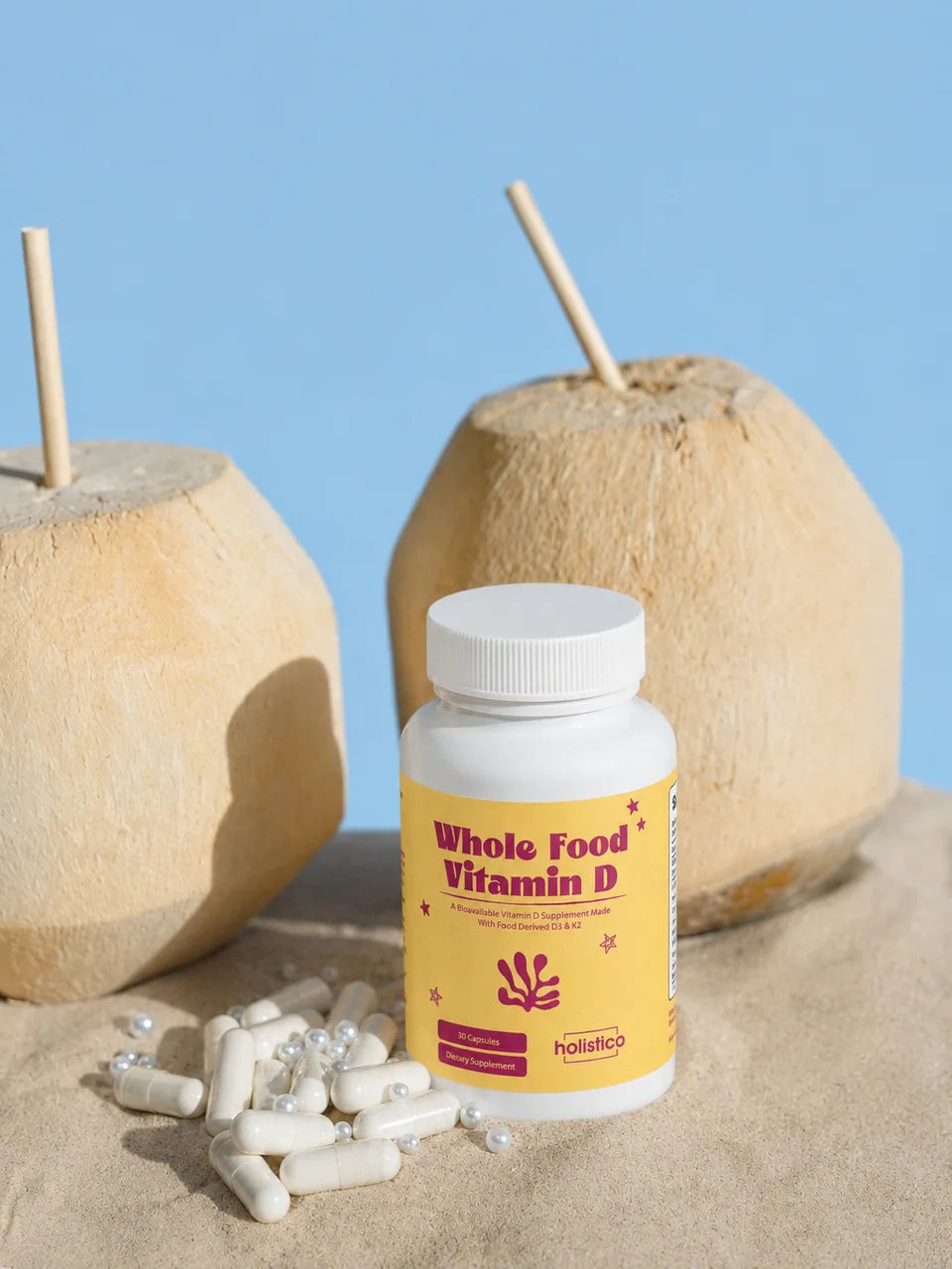 A bottle of Whole Food Vitamin D in sand between two coconuts with straws, capsules and pearls scattered in front