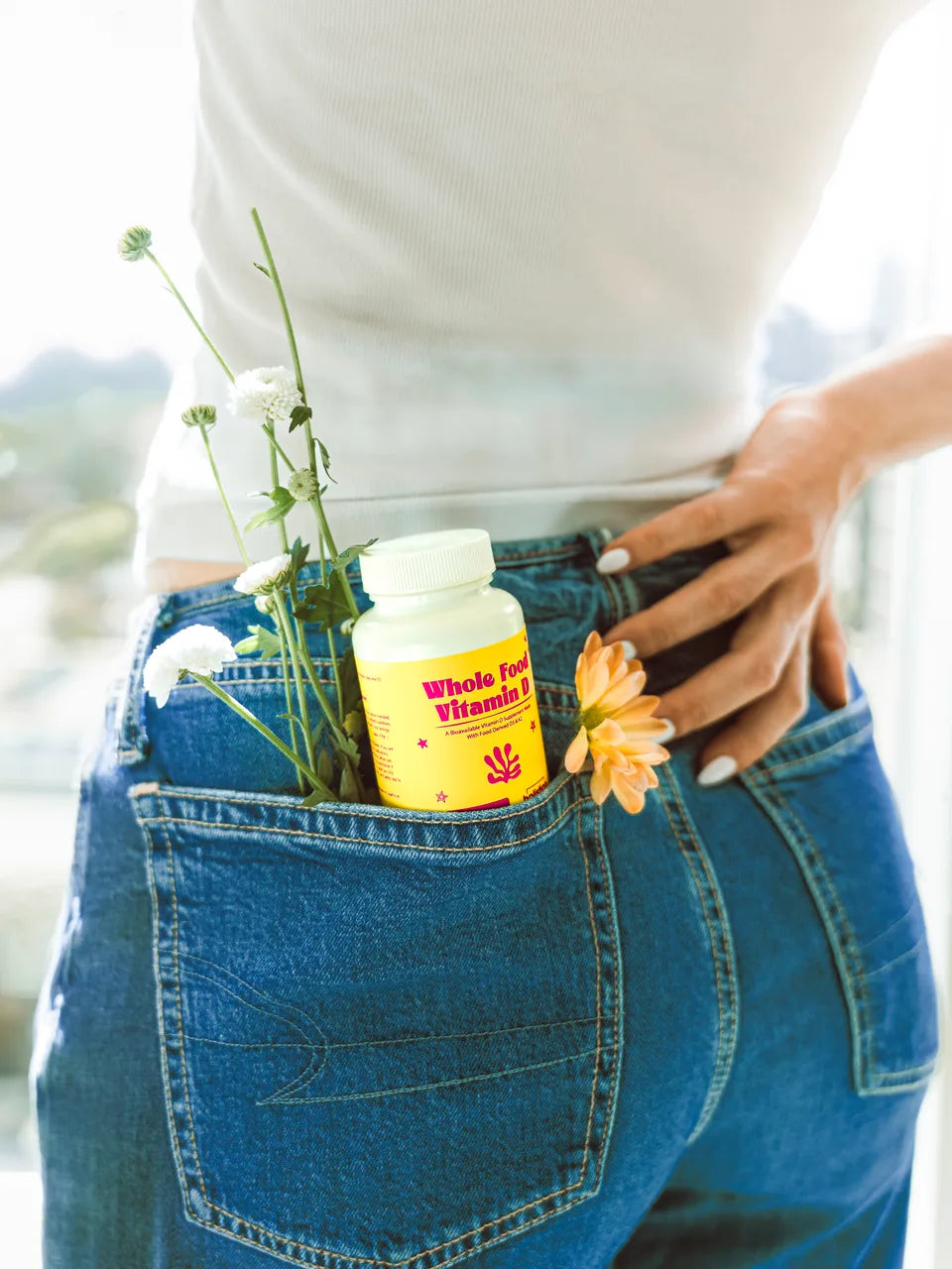 A bottle of Whole Food Vitamin D tucked into the back pocket of blue jeans with a few wildflowers