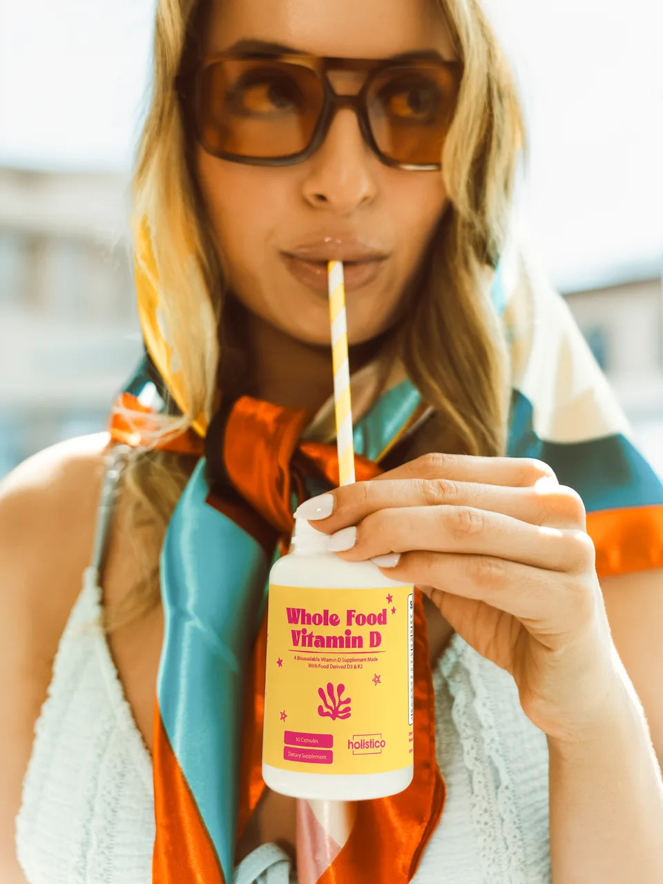 A woman in sunglasses and a silk scarf sipping through a straw from a bottle of Whole Food Vitamin D